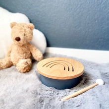 Load image into Gallery viewer, Solid beechwood drum with rainbow design cut into top. Base is a soft dusty rose color. Includes one beechwood mallet with a white head. Drum is placed on a grey fur rug against a deep sea blue background with a brown teddy bear and white plush cloud.